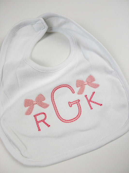 White cotton bib embroidered with a custom pink monogram and embellished in bows.