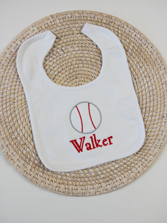 White cotton baby bib embroidered with a baseball design and personalized with name in red thread. Velcro closure.