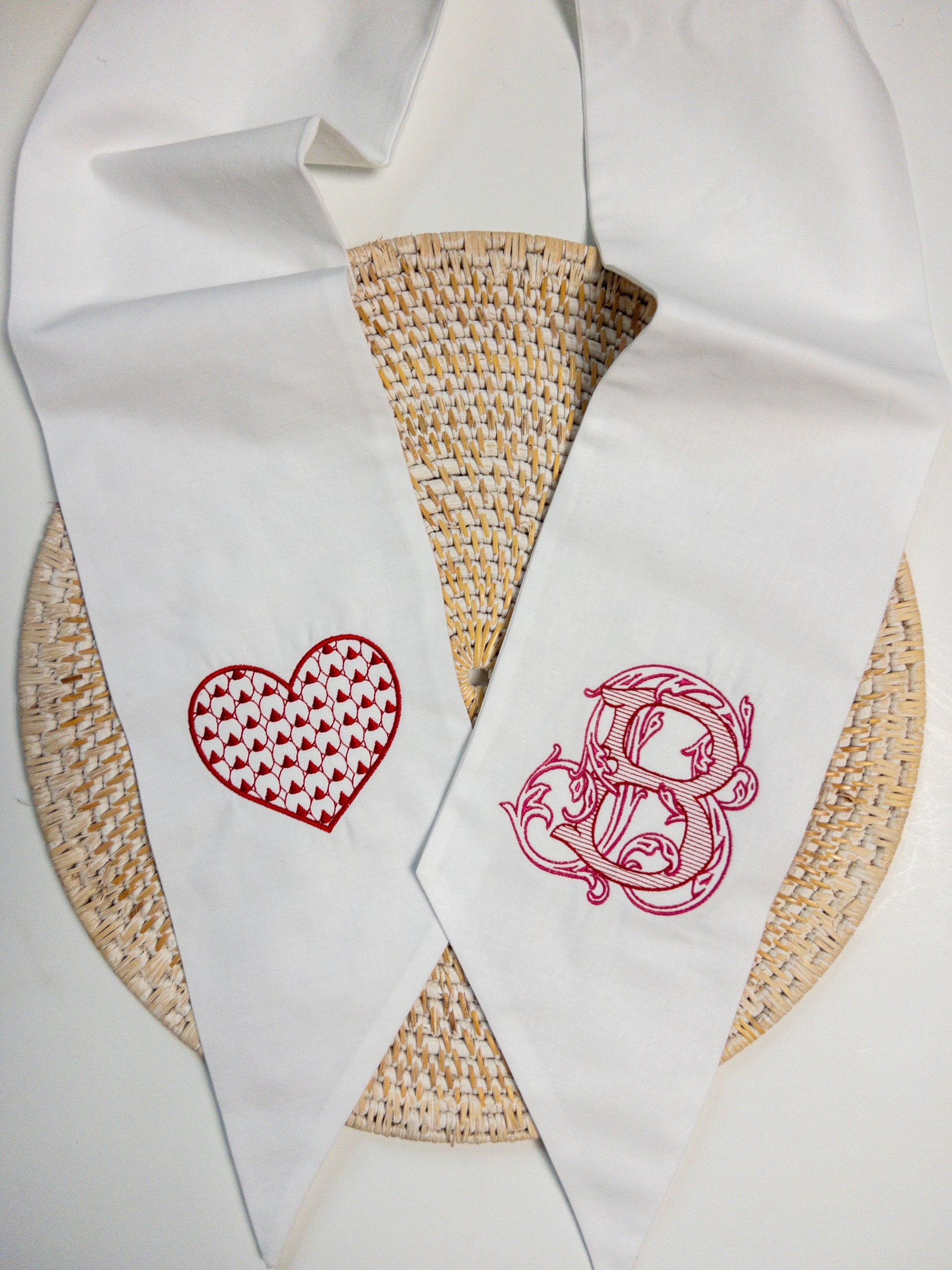 White wreath sash embroidered with a red heart on one end and a fancy monogram on the other side in pink and red thread.